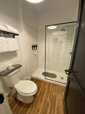 A bathroom with a toilet, a shower with a glass door, white tiled walls, and a wooden floor.