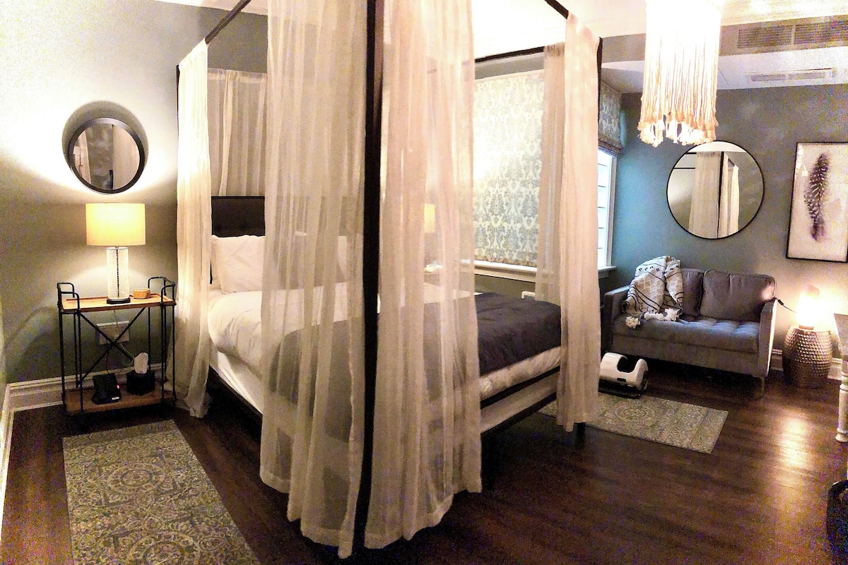 A bedroom with a four-poster bed, a bedside table with a lamp, a small rug, a sofa, and a window with curtains.
