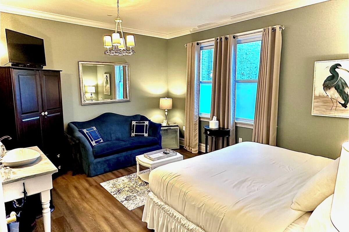 A hotel room with a bed, a blue sofa, a desk with a mirror, a chandelier, and a window with curtains.