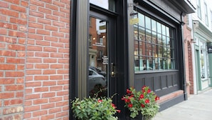 A hotel entrance with a black door, a brick facade, and red flowers in front.