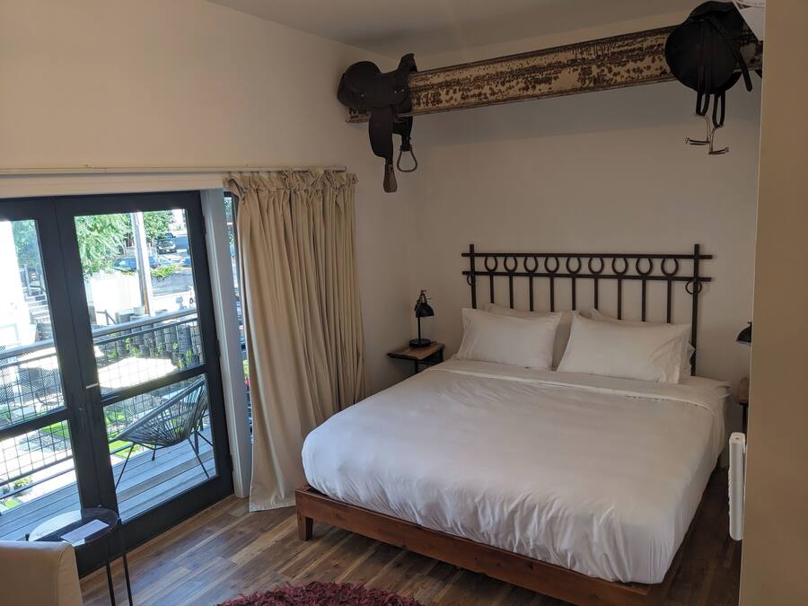 Un dormitorio con cama de madera, una mesita de noche, una lámpara y vistas al exterior a través de un amplio ventanal con cortinas.