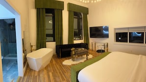 A modern bedroom with a large bed, a sofa, a glass coffee table, a TV, and a shower area.