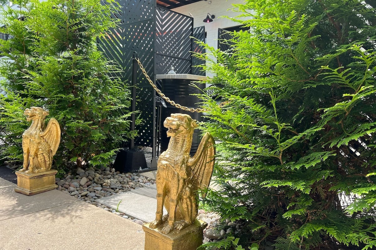 Two golden statues of winged lions in a garden with dense green foliage and a black metal fence in the background.