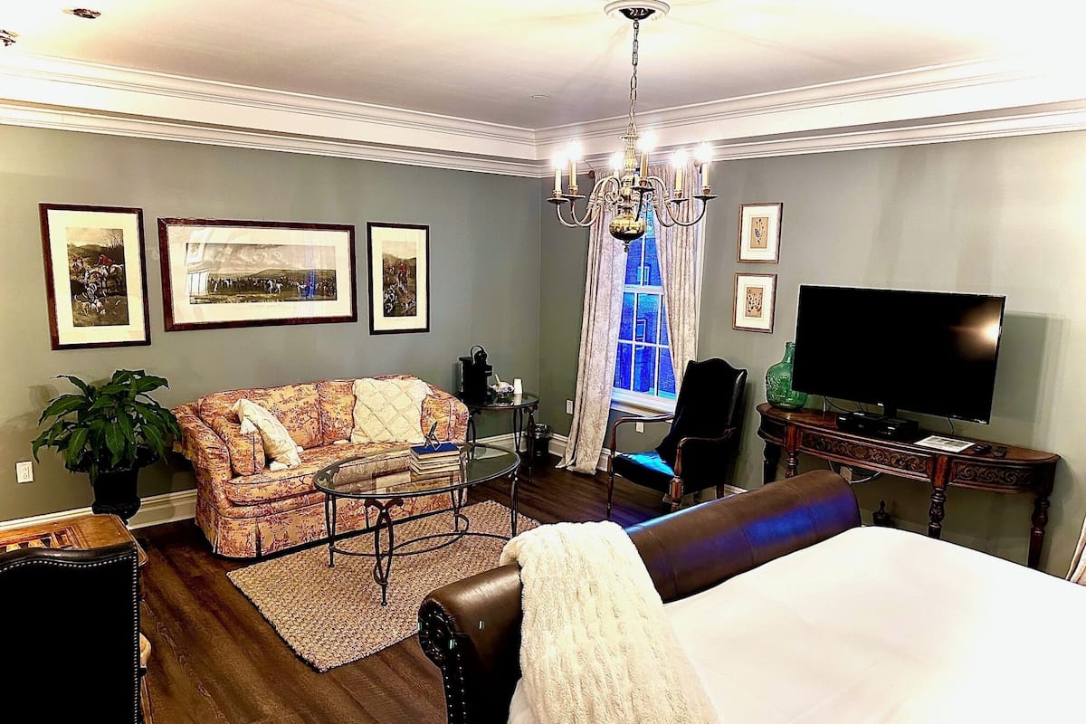 A hotel room with a bed, a sofa, a television, a chandelier, and framed pictures on the wall.