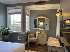 A bedroom with a large window, a bedside chair, a vanity with a mirror, and a bed.