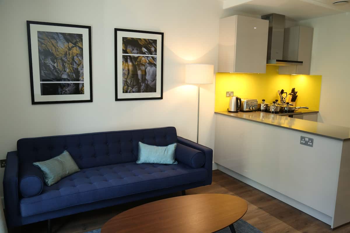 A modern living room with a dark blue sofa, two framed abstract paintings, a floor lamp, and a kitchen area with a yellow backsplash and white cabinets.