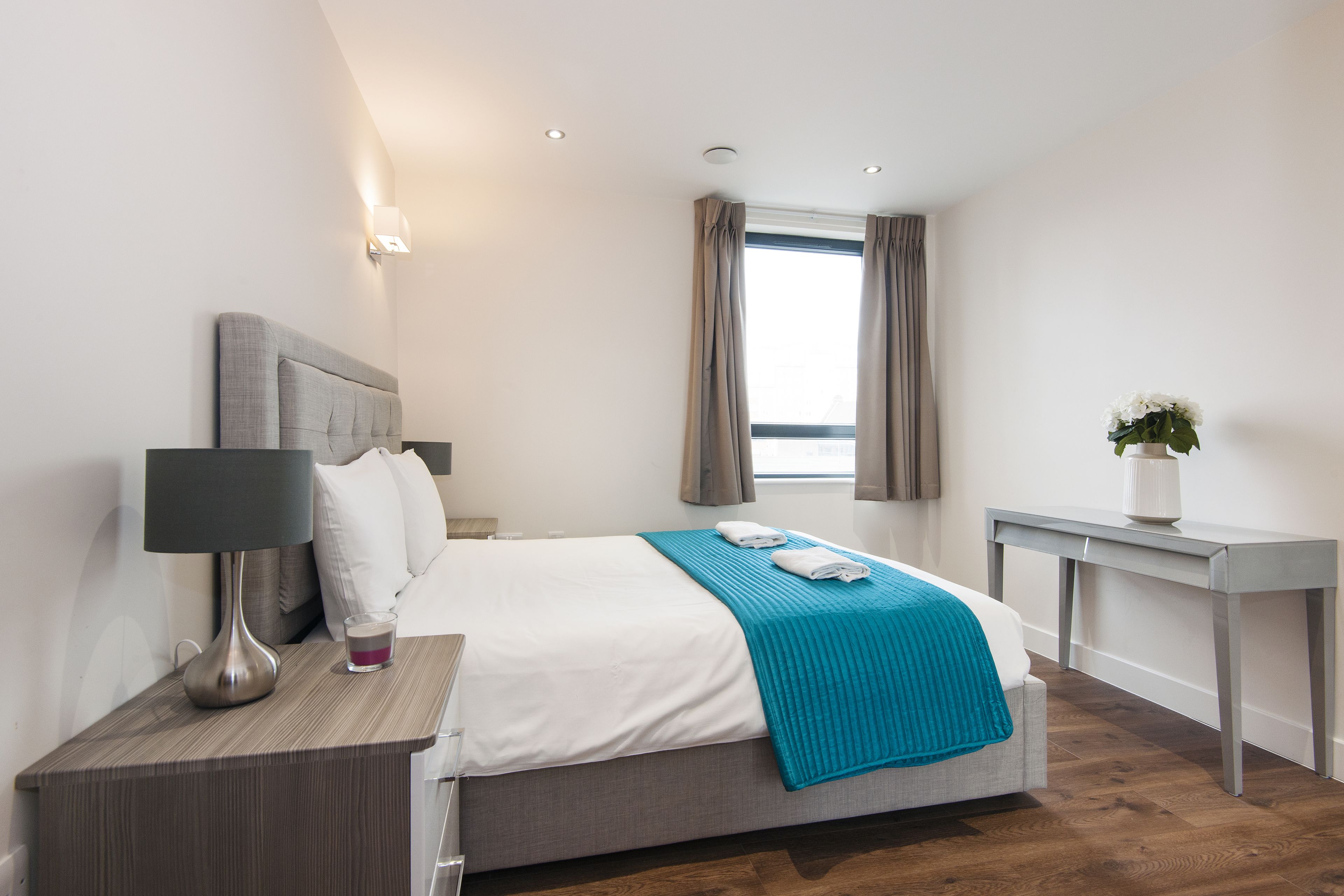 A modern hotel room with a neatly made bed, a bedside table with a lamp, a glass of water, and a view of the outdoors through a window with curtains.