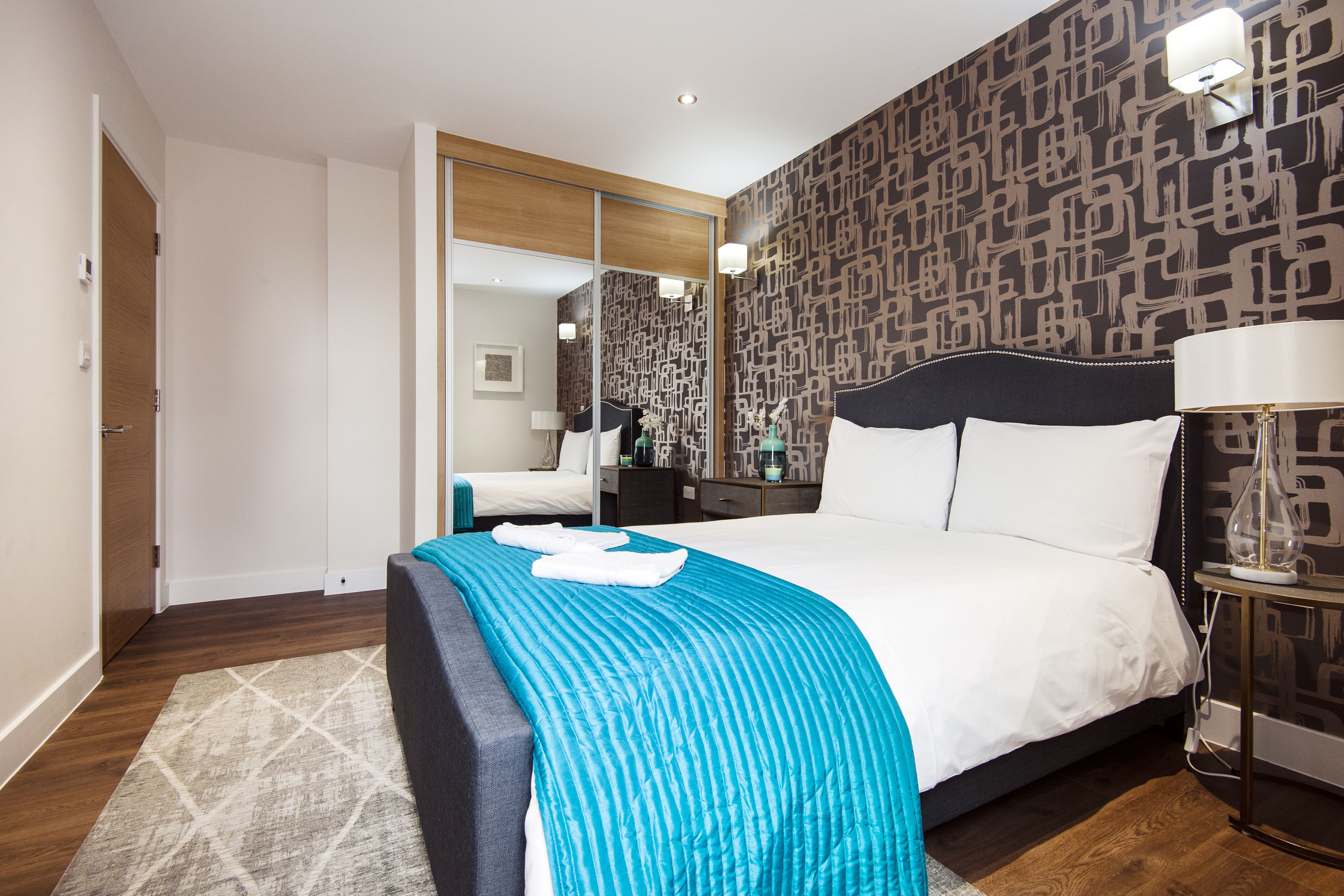 Habitación de hotel moderna con una cama grande, un espejo, una mesita de noche con lámpara y una pared con diseño.