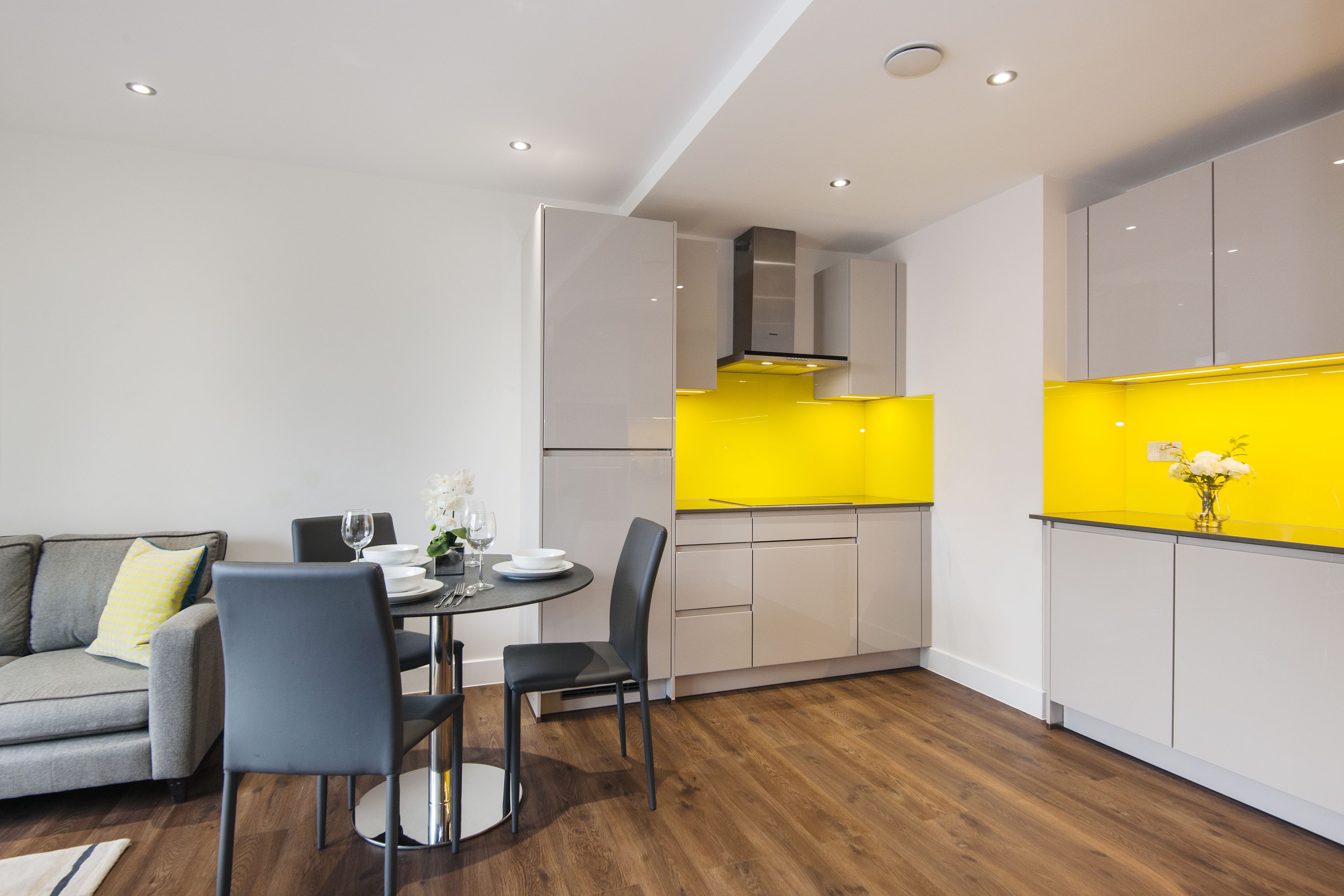 Eine moderne Küche mit weißen Schränken und einer gelben Spritzschutzwand, ein Essbereich mit einem grauen Sofa und einem runden Tisch, sowie Holzböden.