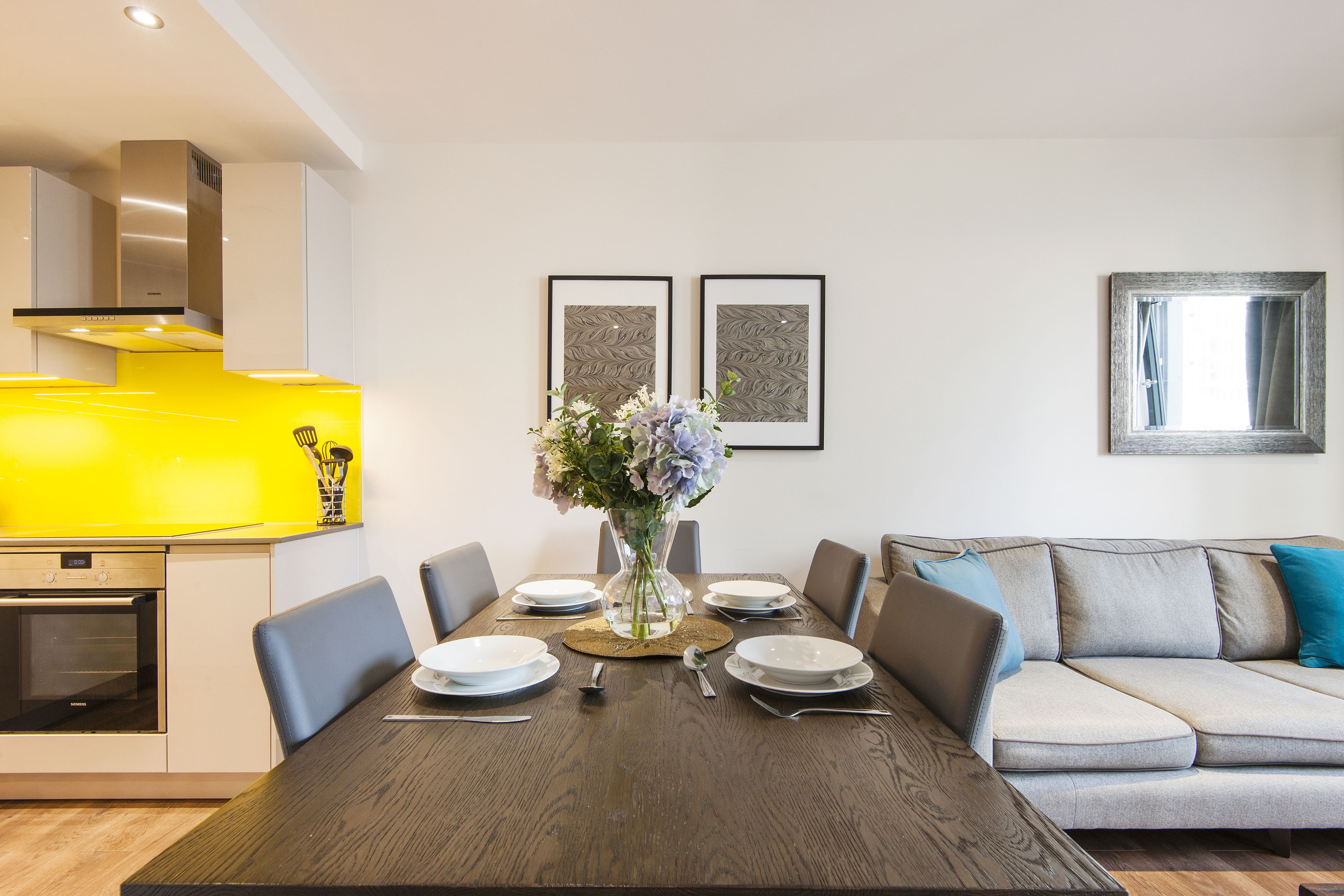 Et moderne spiseområde med et træbord, stole og en vase med blomster. Der er en sofa og et vindue med gardiner.