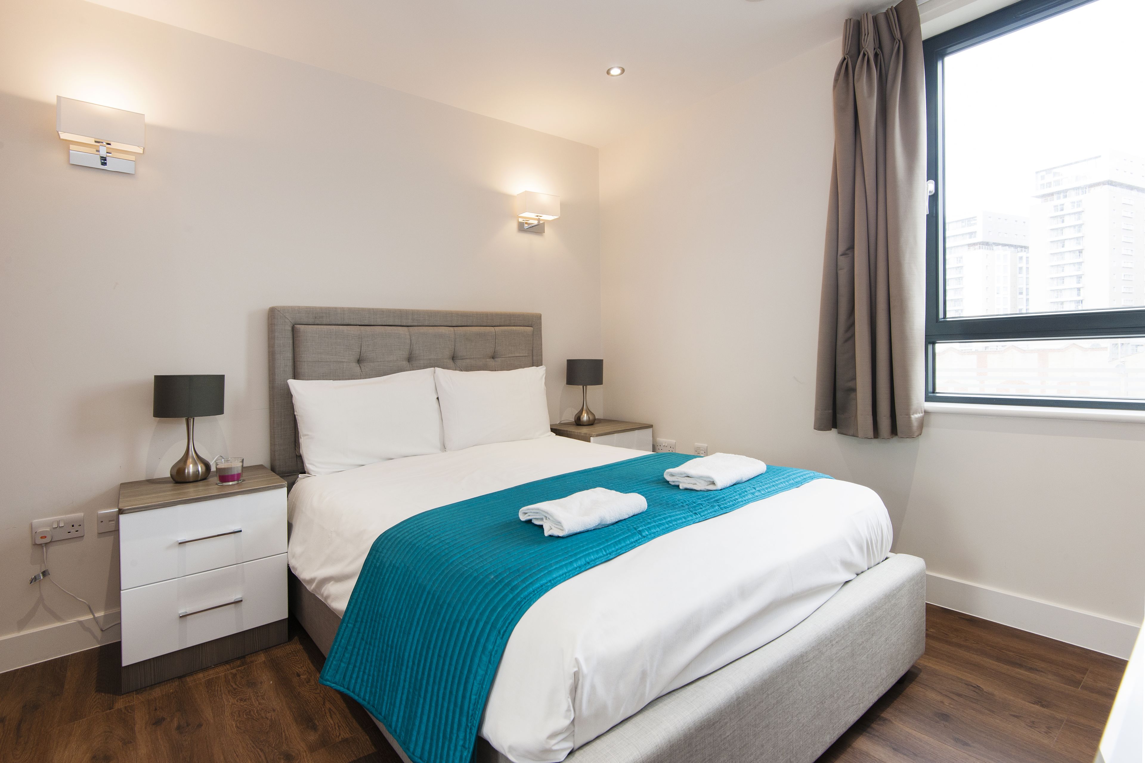 A hotel room with a bed, nightstands, a lamp, a window, and a view of buildings.