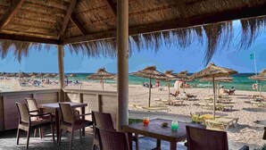 Un espace extérieur aménagé avec des chaises et des tables en osier, donnant sur une plage parsemée de parasols et fréquentée par des gens.