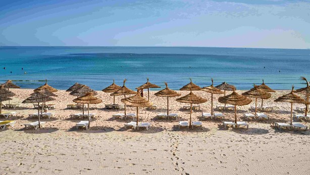 Une plage bordée de rangées de chaises longues et d’ombrelles en chaume, un ciel bleu dégagé et une mer calme.