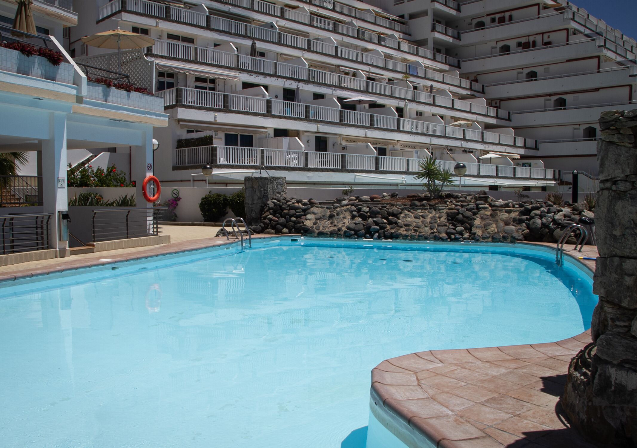 A swimming pool with a stone wall and a multi-story building in the background.