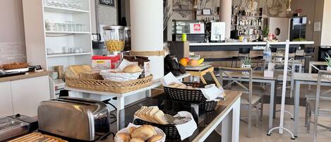 Zona de bufé para el desayuno con diversas opciones de comida, una máquina de café y disposición de mesas.