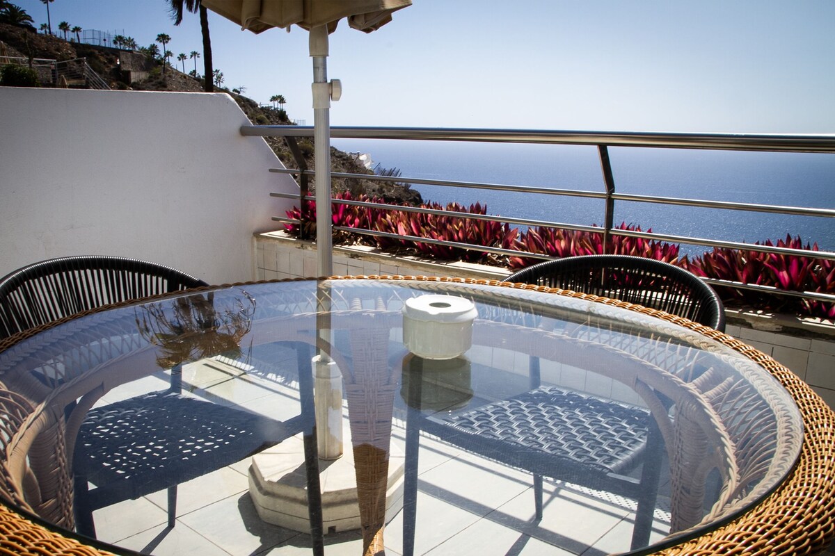 A round glass table with wicker chairs, a white trash bin, and a view of the ocean and red flowers.