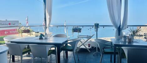 En terrasse med borde og stole med udsigt over en strand, et skilt med 'Holiday Club' og et reception område.