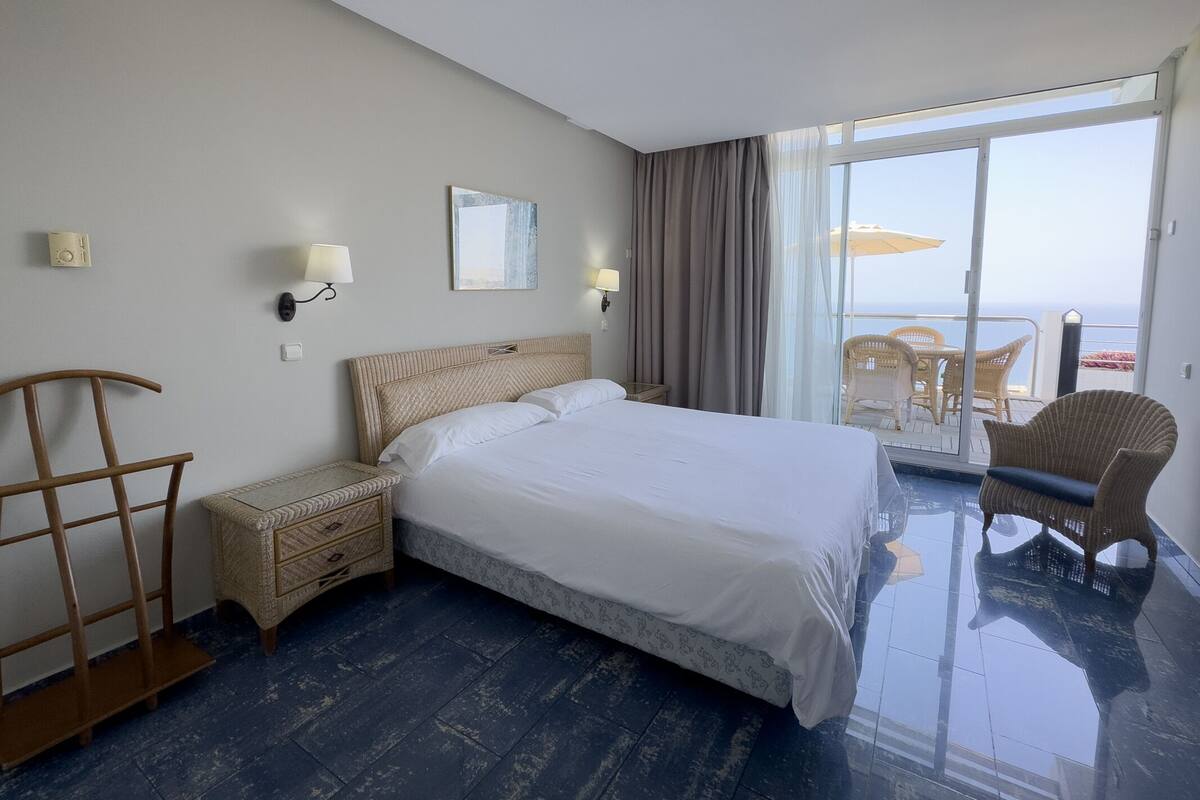 Hotelkamer met een groot bed, een nachtkastje, een stoel en uitzicht op de oceaan door een schuifdeur.