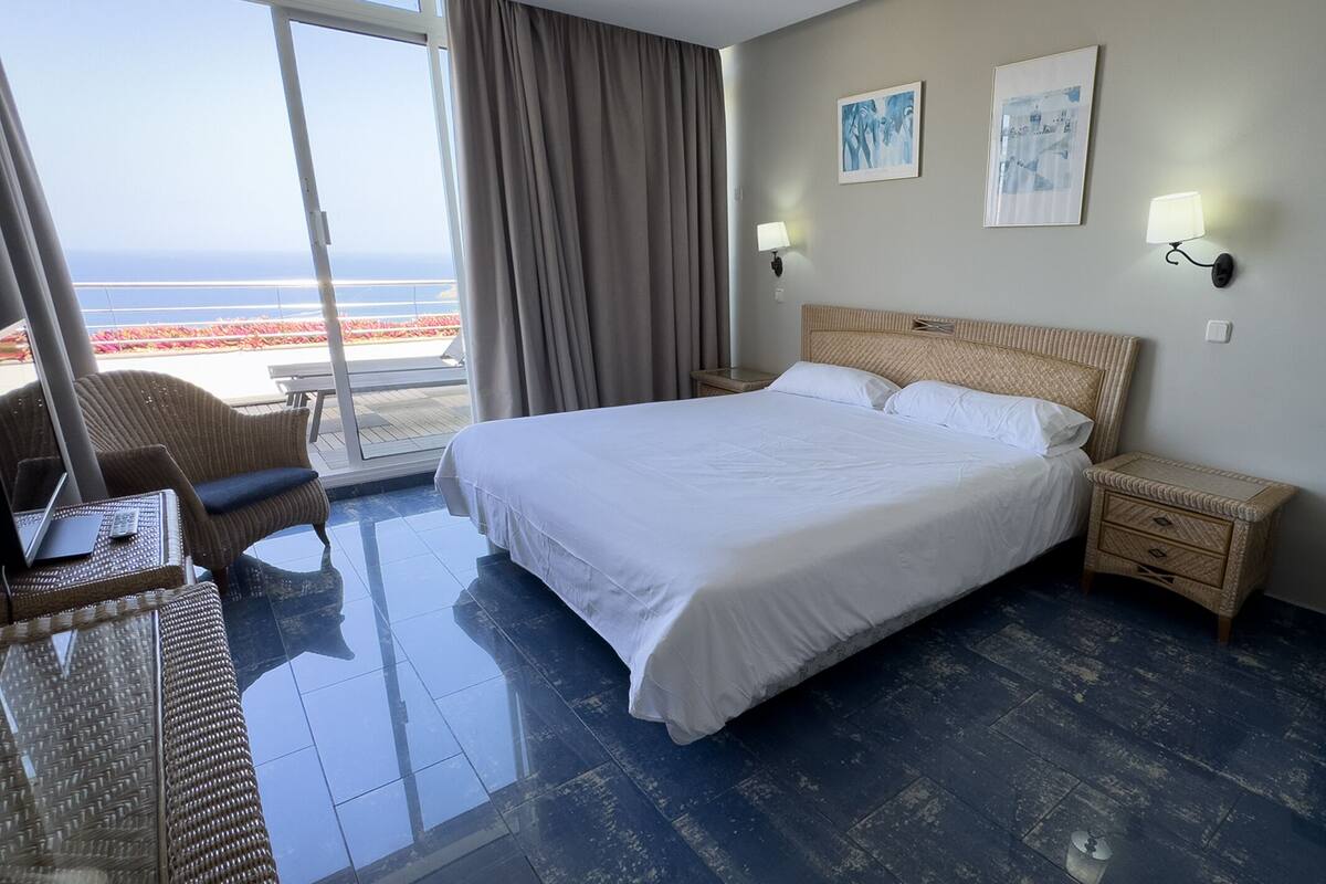 Een hotelkamer met een groot bed, een stoel, een tafeltje en uitzicht op het strand.