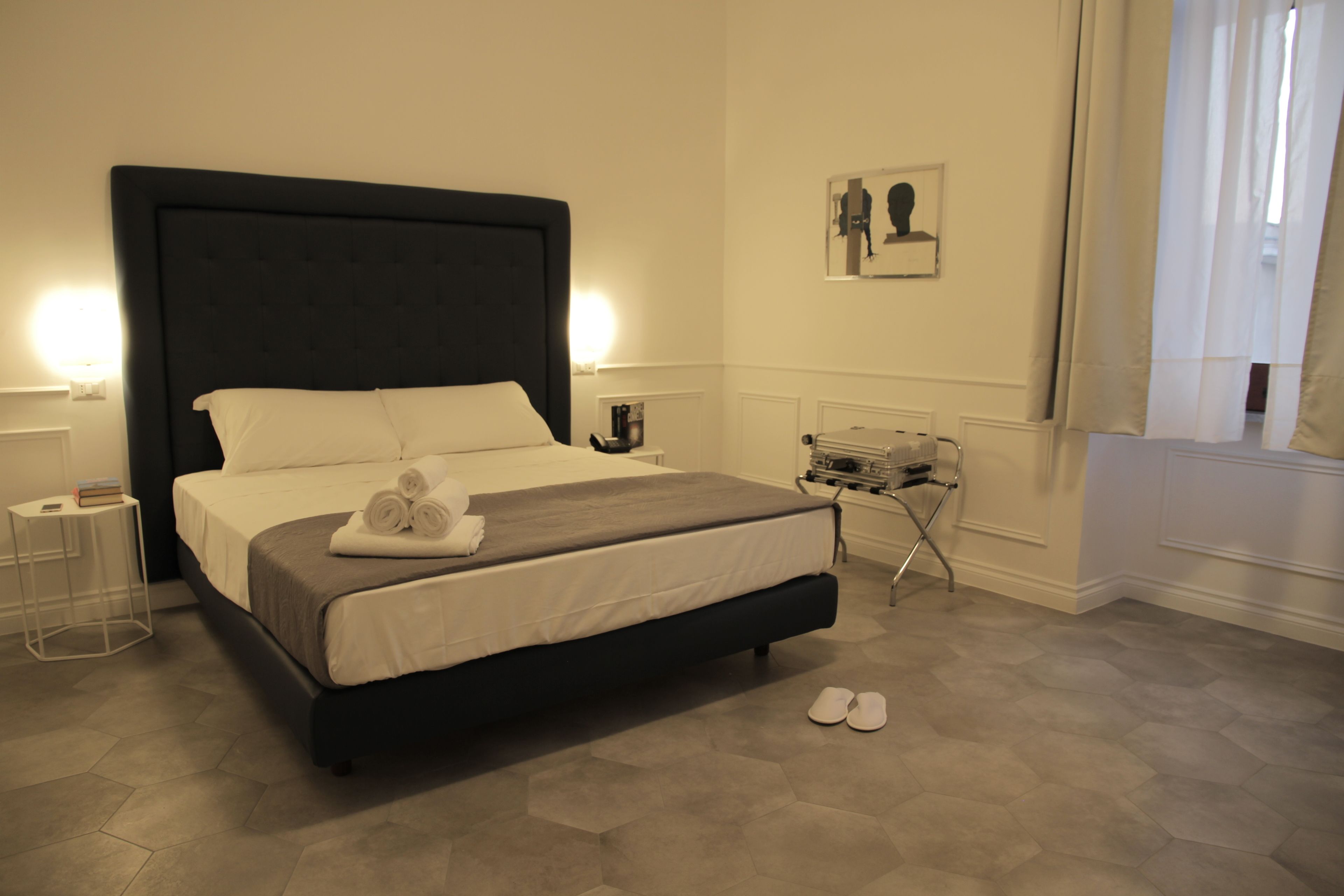 Cama bem arrumada com cabeceira escura, mesa de cabeceira com abajur e uma pequena mesa com um laptop.