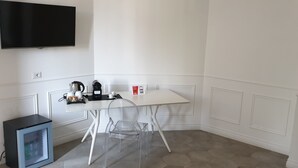 Een kleine, witte kamer met een witte tafel, een transparante stoel, een kleine zwarte koelkast en een flatscreen-televisie aan de muur.