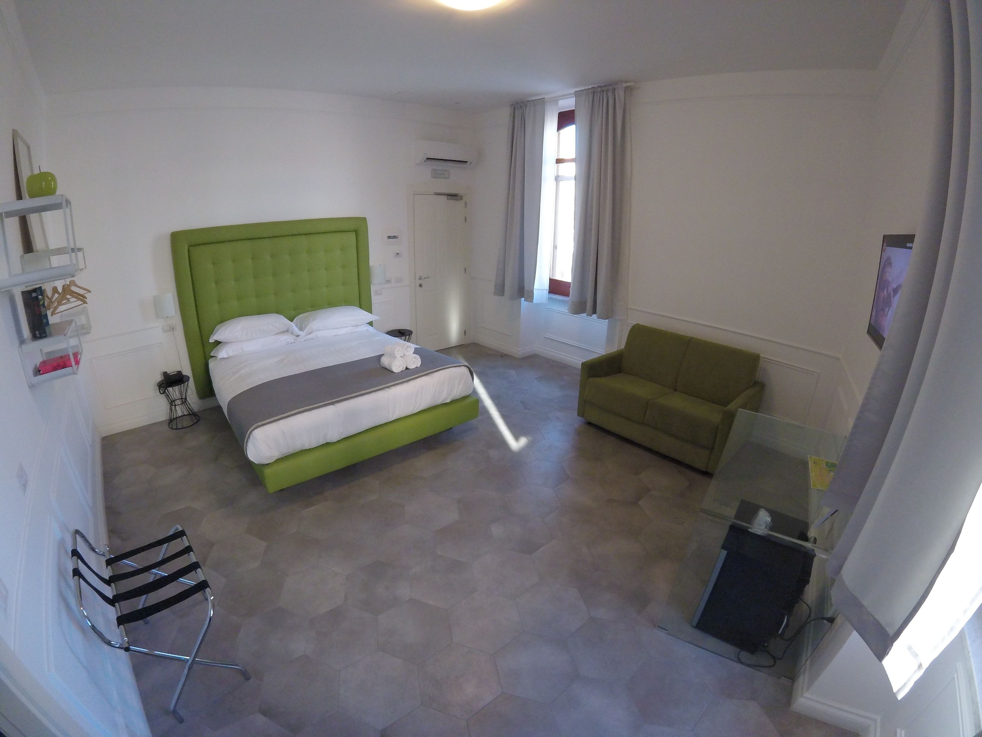 Quarto de hotel com uma cama estofada em verde, uma poltrona verde, uma mesa pequena e uma cadeira.