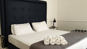 Een net opgemaakt bed met witte lakens en een donker hoofdbord, een nachtkastje met een lamp, en een stapje opgerolde handdoeken op het bed.