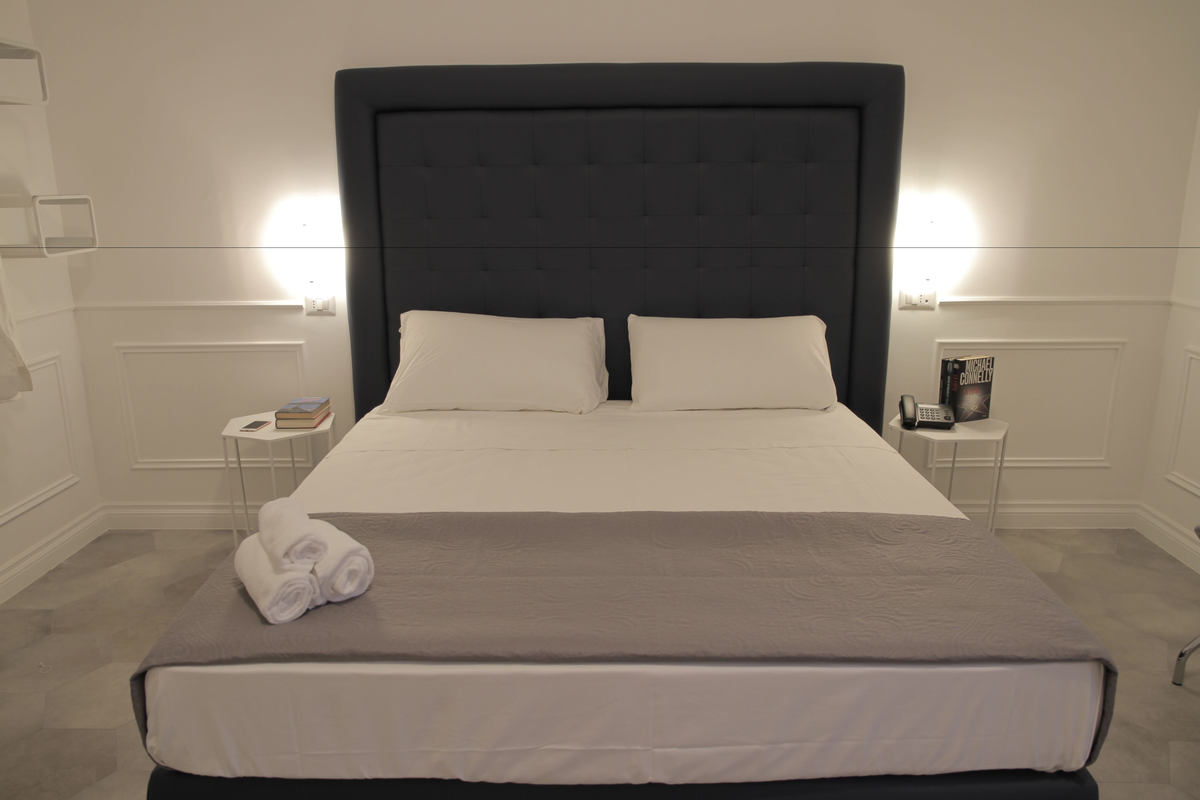 A neatly made bed with a dark headboard, white pillows, and a folded towel on the footboard. There are two bedside tables with a book and a phone on one, and a vase on the other. The room has a clean, minimalist design with white walls and a light-colored floor.