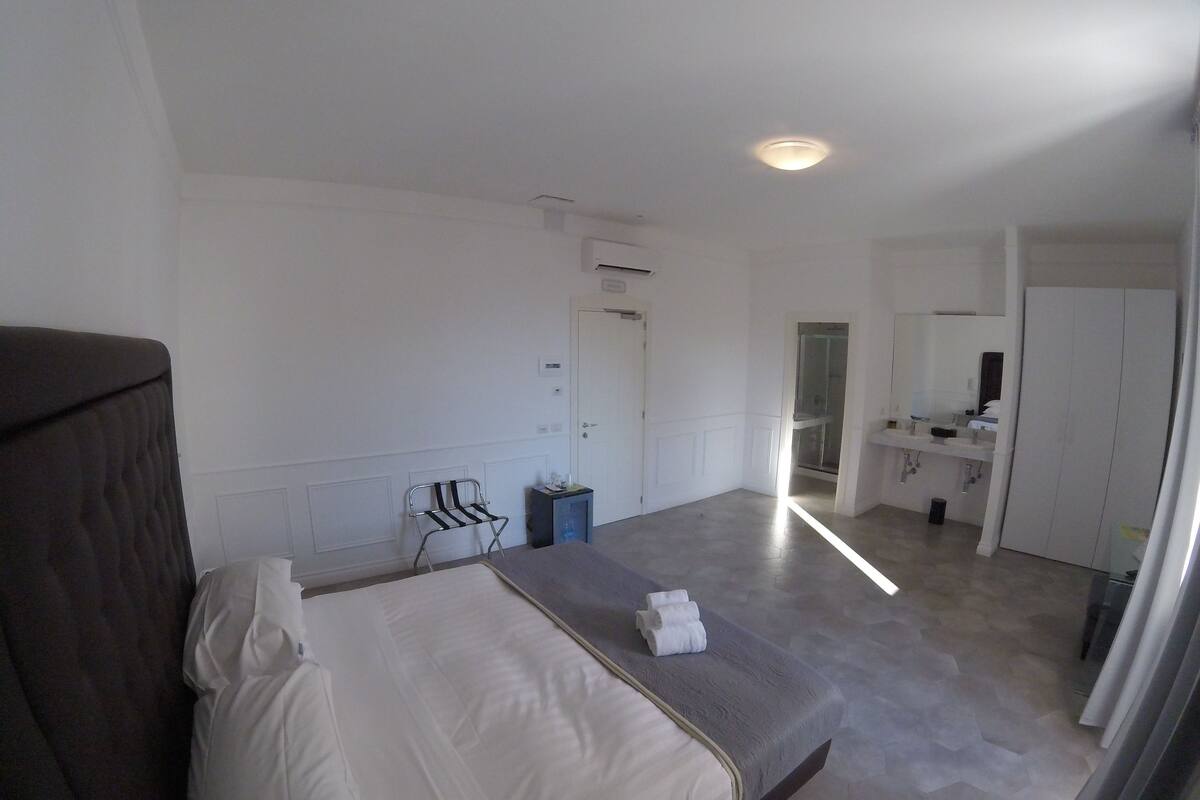 Une chambre d’hôtel comprenant un lit, une chaise, une petite table, une salle de bain avec un lavabo et un miroir, et une porte donnant sur une autre pièce.