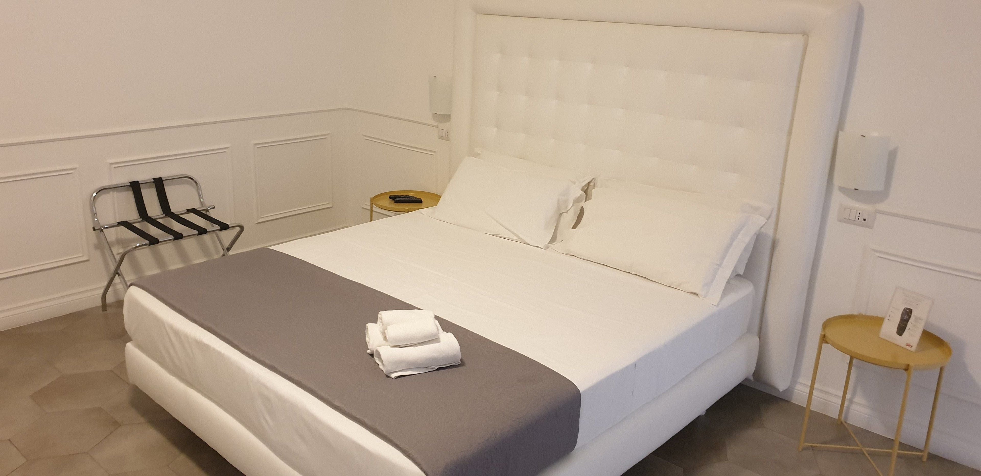 Ein ordentlich gemachtes Bett mit weißen Bettwäsche und grauem Akzent, ein kleiner Nachttisch mit Fernbedienung sowie ein an der Wand befestigter Handtuchhalter.
