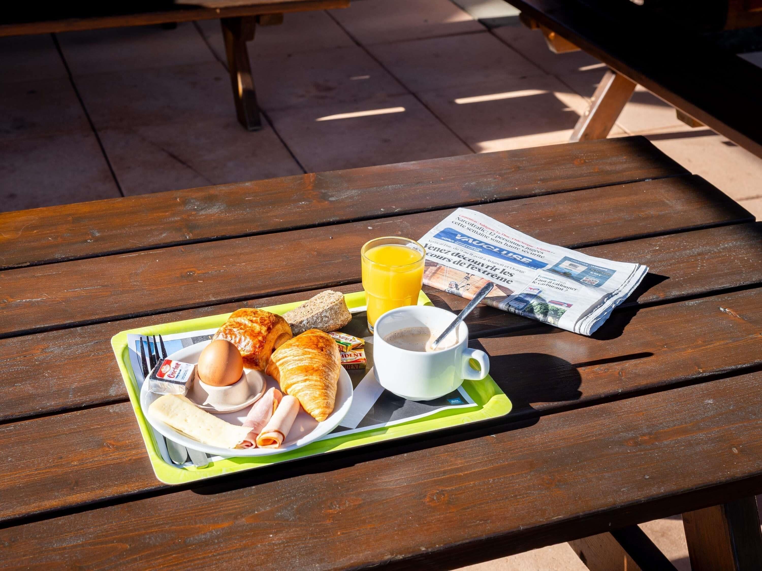 Uma mesa de piquenique com uma bandeja contendo croissants, um sanduíche, um ovo cozido, um copo de suco de laranja e uma xícara de café.