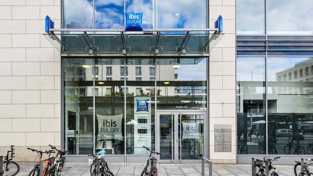 A modern hotel with a glass facade, a prominent 'ibis budget' sign, and bicycles parked outside.