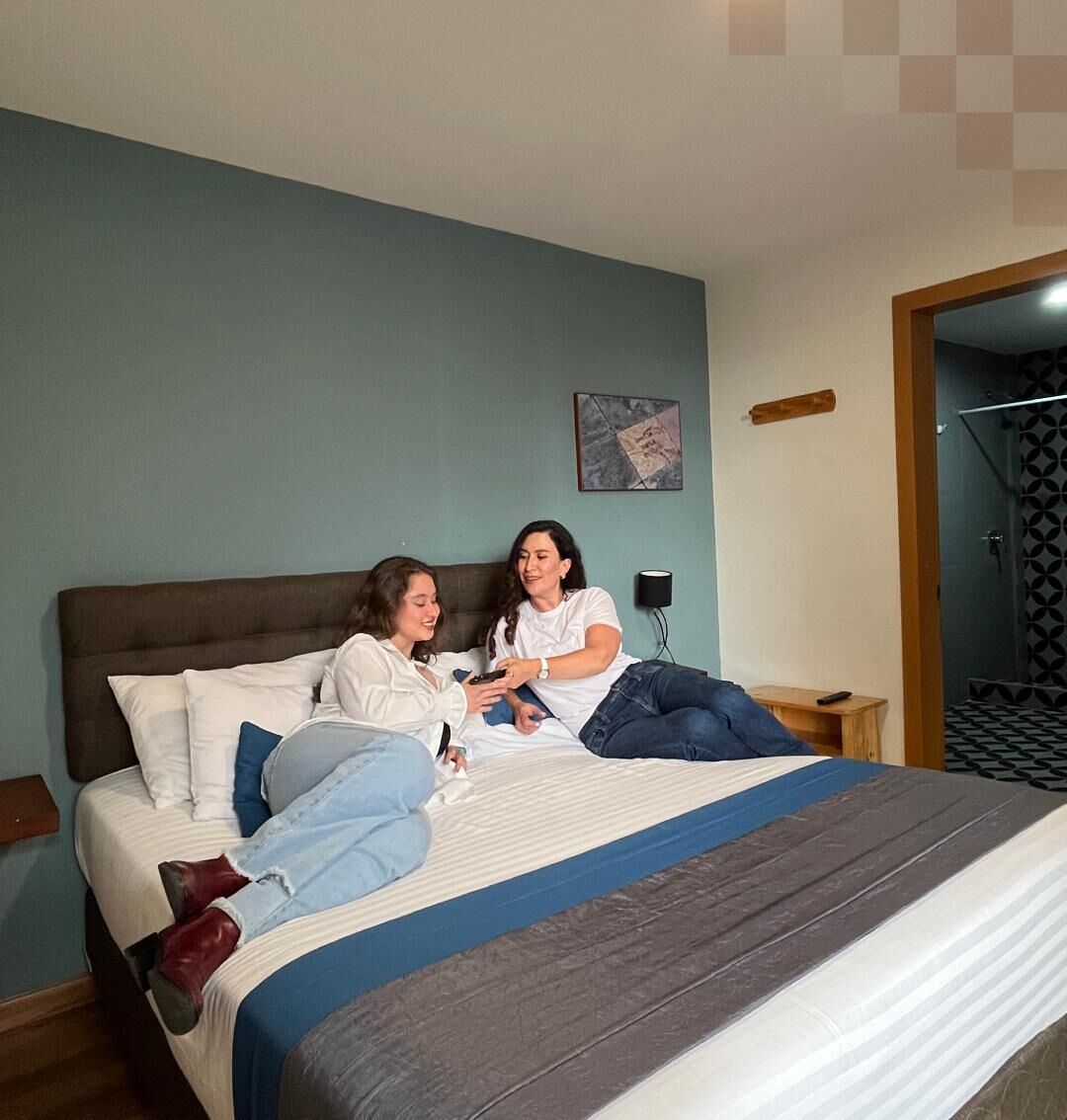 A bedroom with a bed, two people sitting on it, a nightstand, and a wall with a picture.