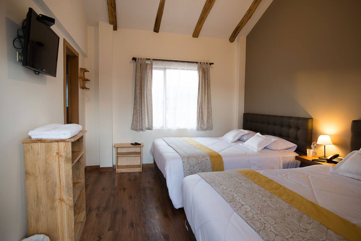 Habitación de hotel con dos camas, un aparador de madera, televisor, ventana con cortinas y una lámpara de noche.