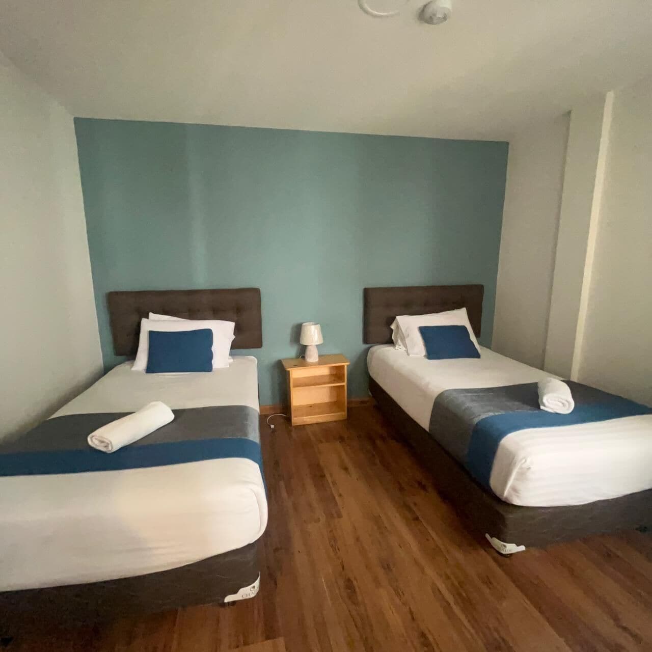 A hotel room with two single beds, each with a headboard, a bedside table, and a lamp. The room has a wooden floor and teal walls.