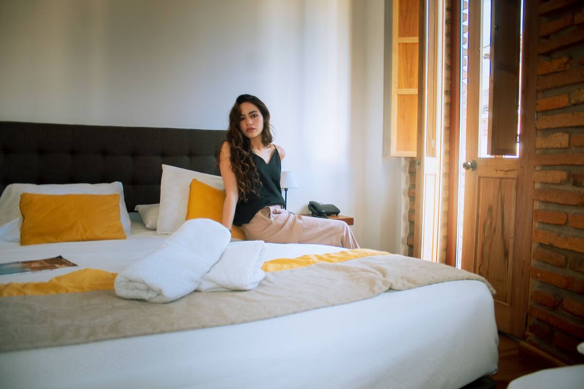 Una cama con una colcha blanca y amarilla, un cabecero negro y una puerta de madera frente a una pared de ladrillo.