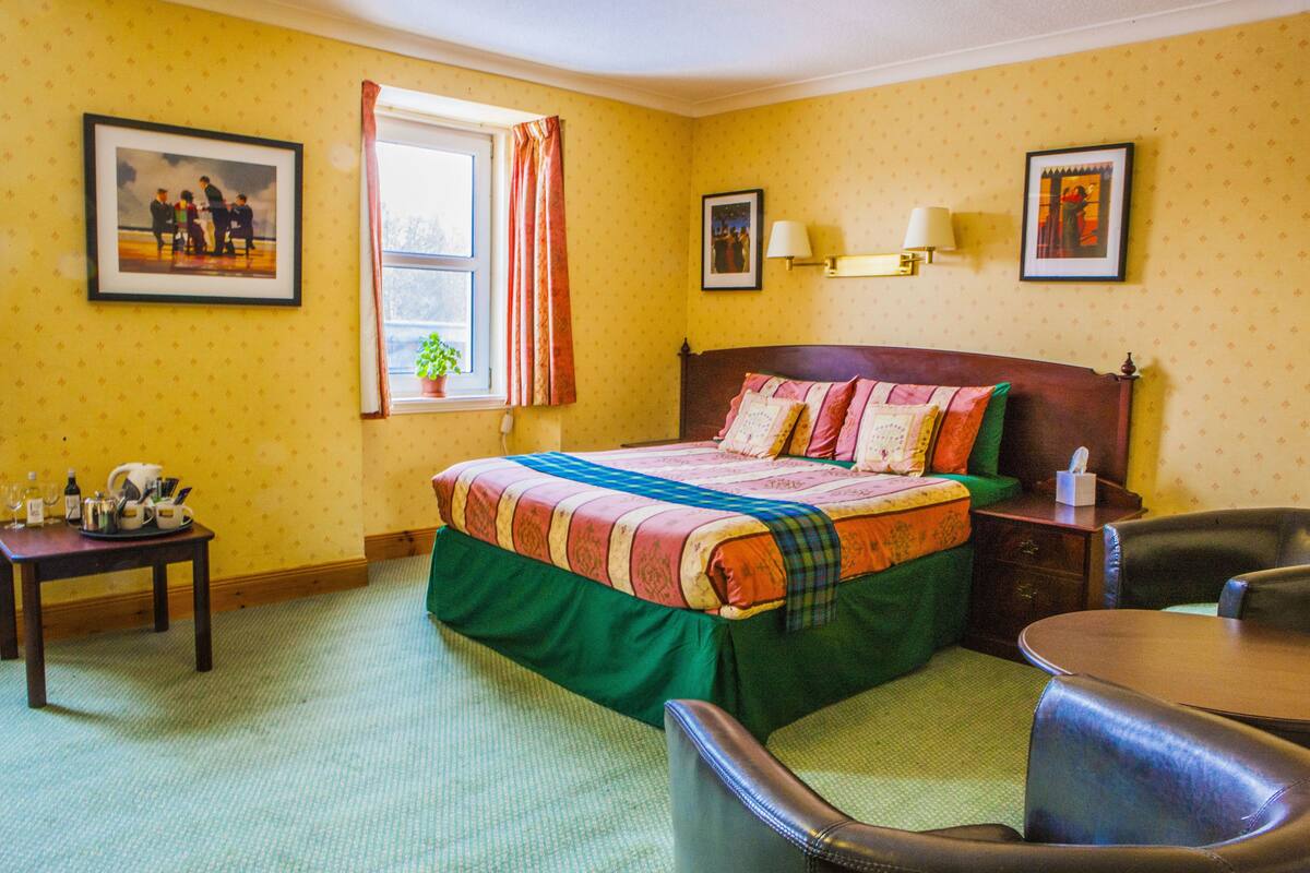 A hotel room with a large bed, a bedside table with a lamp, a chair, and a small table with a plant.