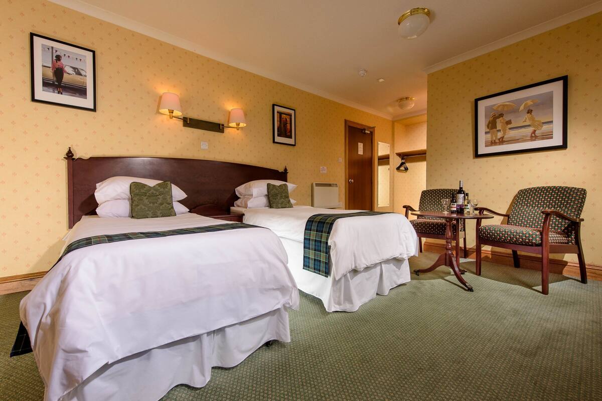 A hotel room with two beds, a small table with a bottle, and a chair.