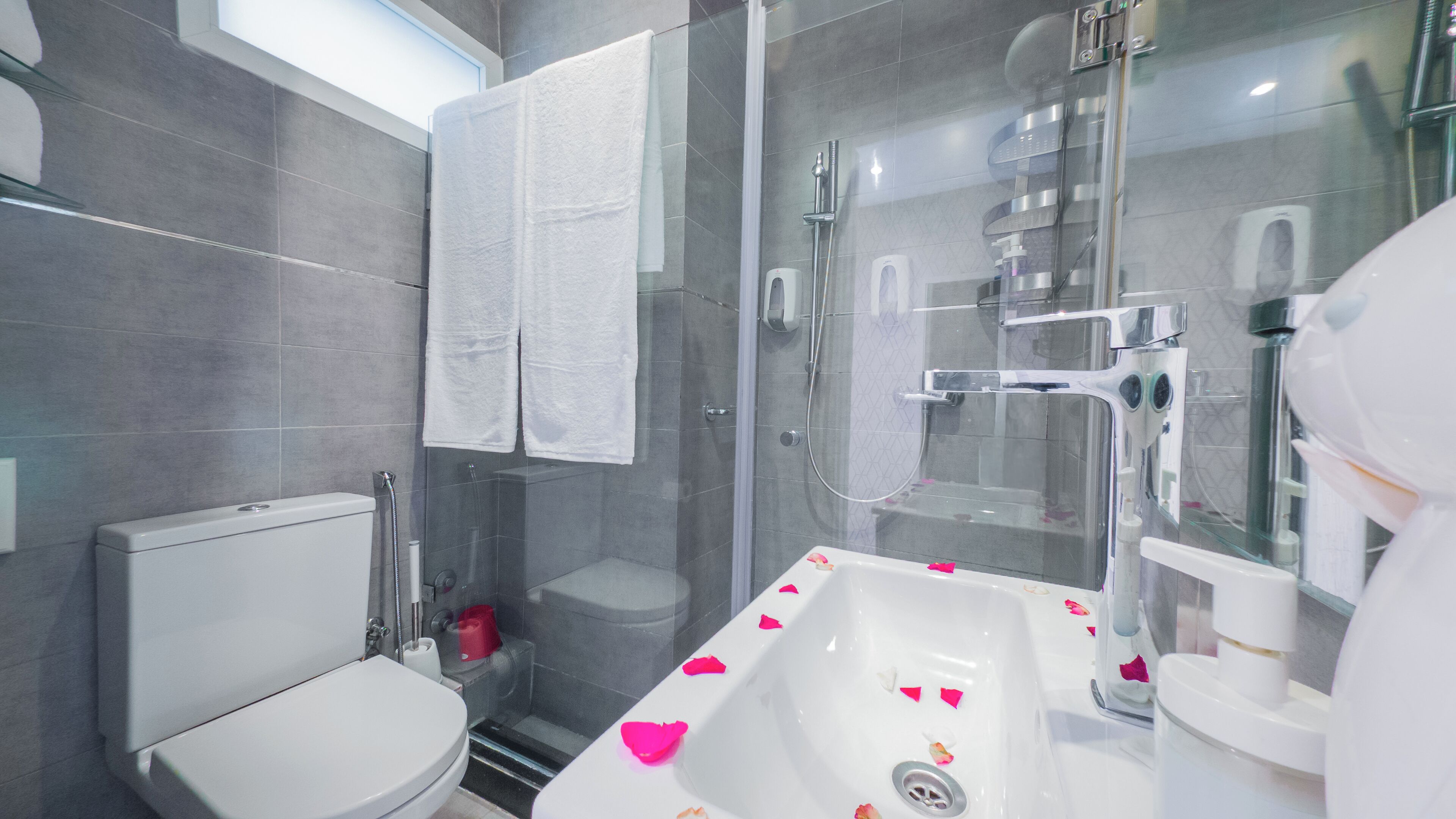 A modern bathroom with a glass-enclosed shower, a white toilet, and a sink with rose petals.