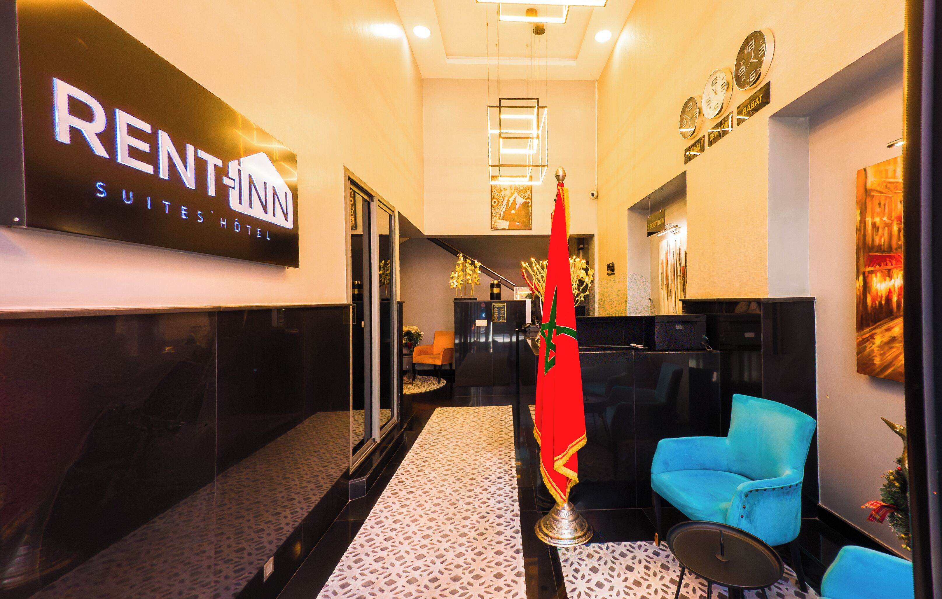 Un hall d'accueil d'hôtel avec une enseigne indiquant « RENT-INN SUITES HOTEL », un comptoir d'accueil, un drapeau rouge et des chaises bleues.