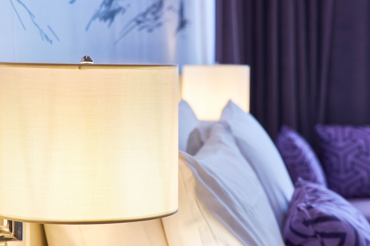 Une lampe avec un abat-jour beige, un lit avec des coussins blancs et violets, et un tableau accroché au mur.