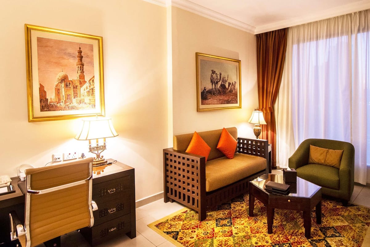 A hotel room with a sofa, armchair, desk, lamps, and two framed pictures on the walls.
