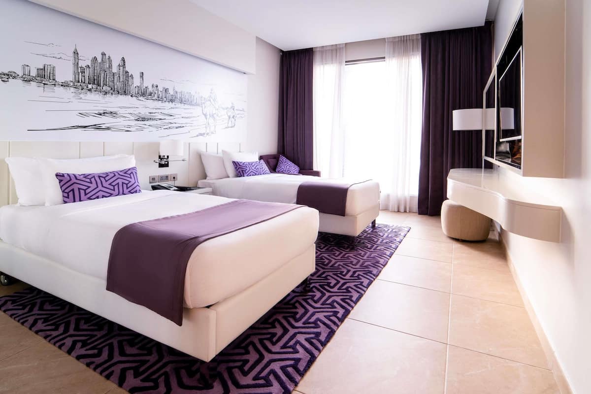 A modern hotel room with two beds, a large cityscape mural, a desk, and a large window with curtains.