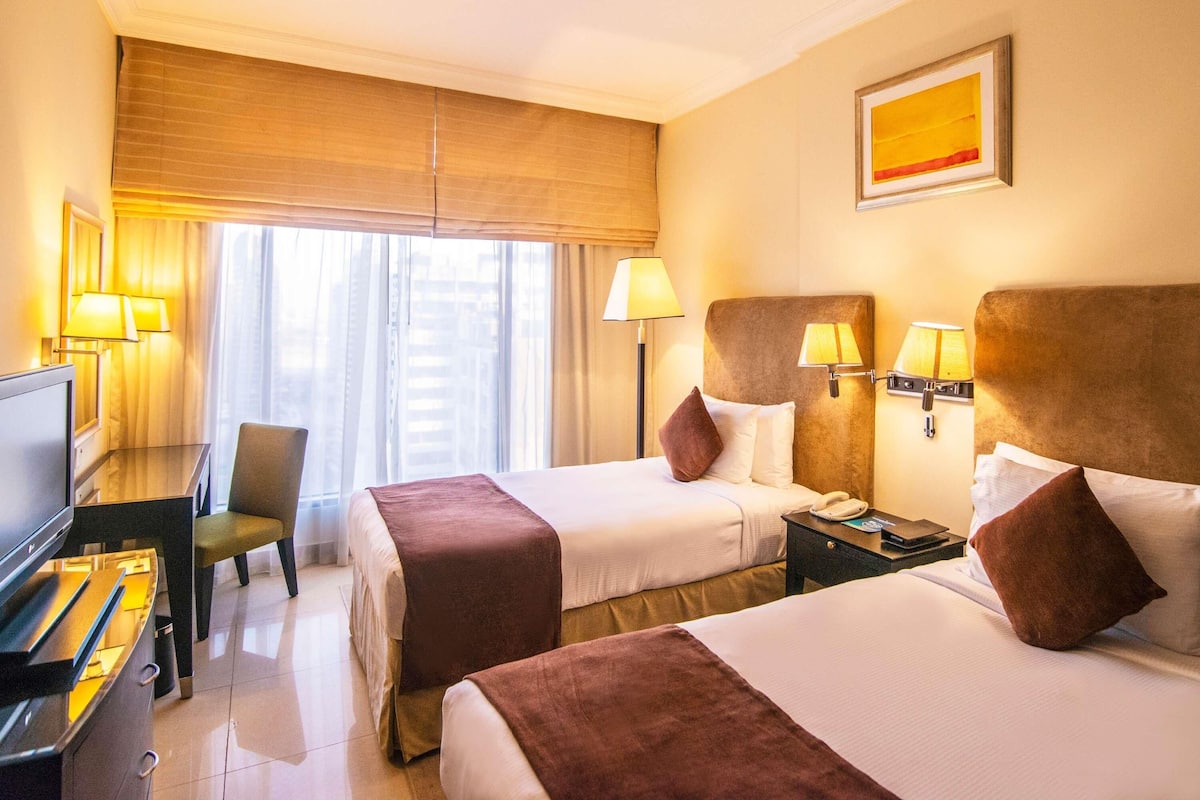 A hotel room with two beds, a desk with a computer, a chair, and a window with blinds.