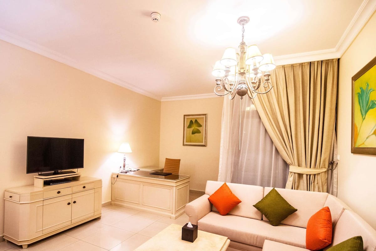A living room with a television, a desk, a sofa with cushions, a chandelier, and a painting on the wall.
