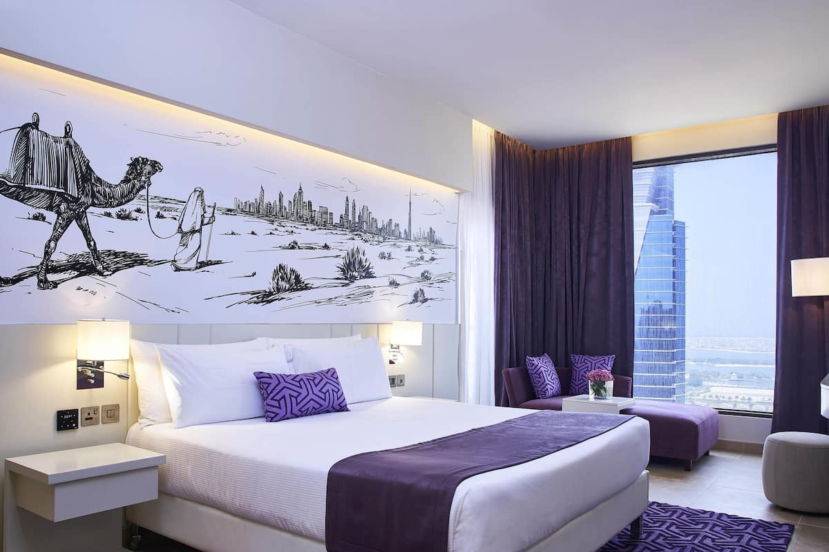 A modern hotel room with a large bed, a nightstand, a seating area with a view of a cityscape, and a window with curtains.
