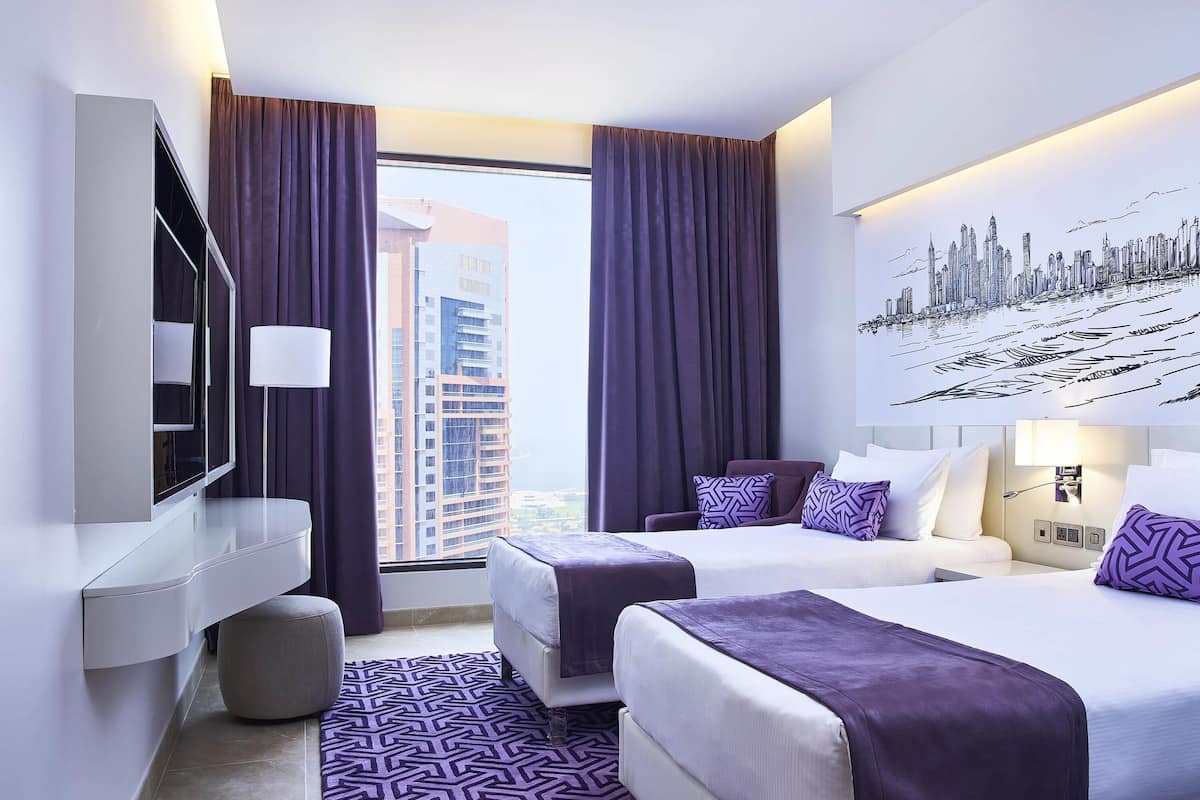 A modern hotel room with two beds, a large window with curtains, a cityscape mural, and a flat-screen TV.