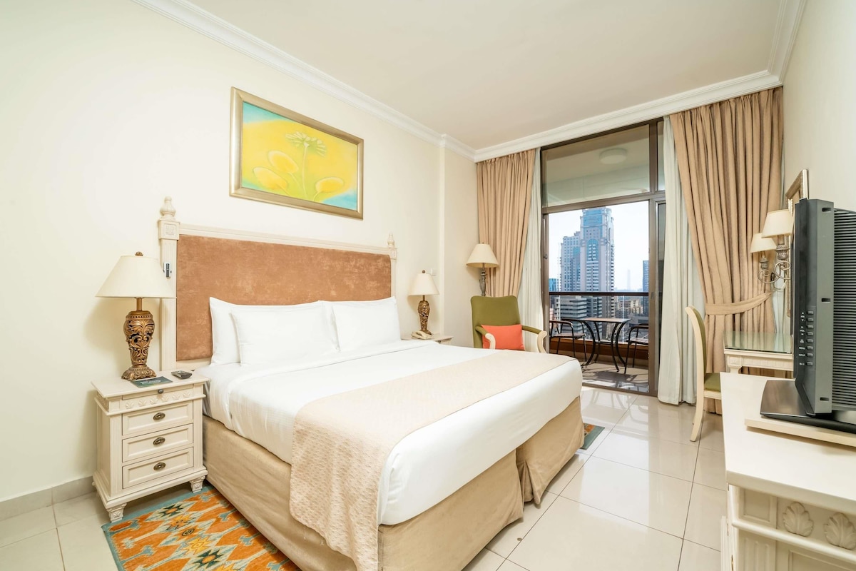 Quarto de hotel com cama, mesas de cabeceira, escrivaninha, cadeira, televisão e vista da cidade através de uma grande janela.