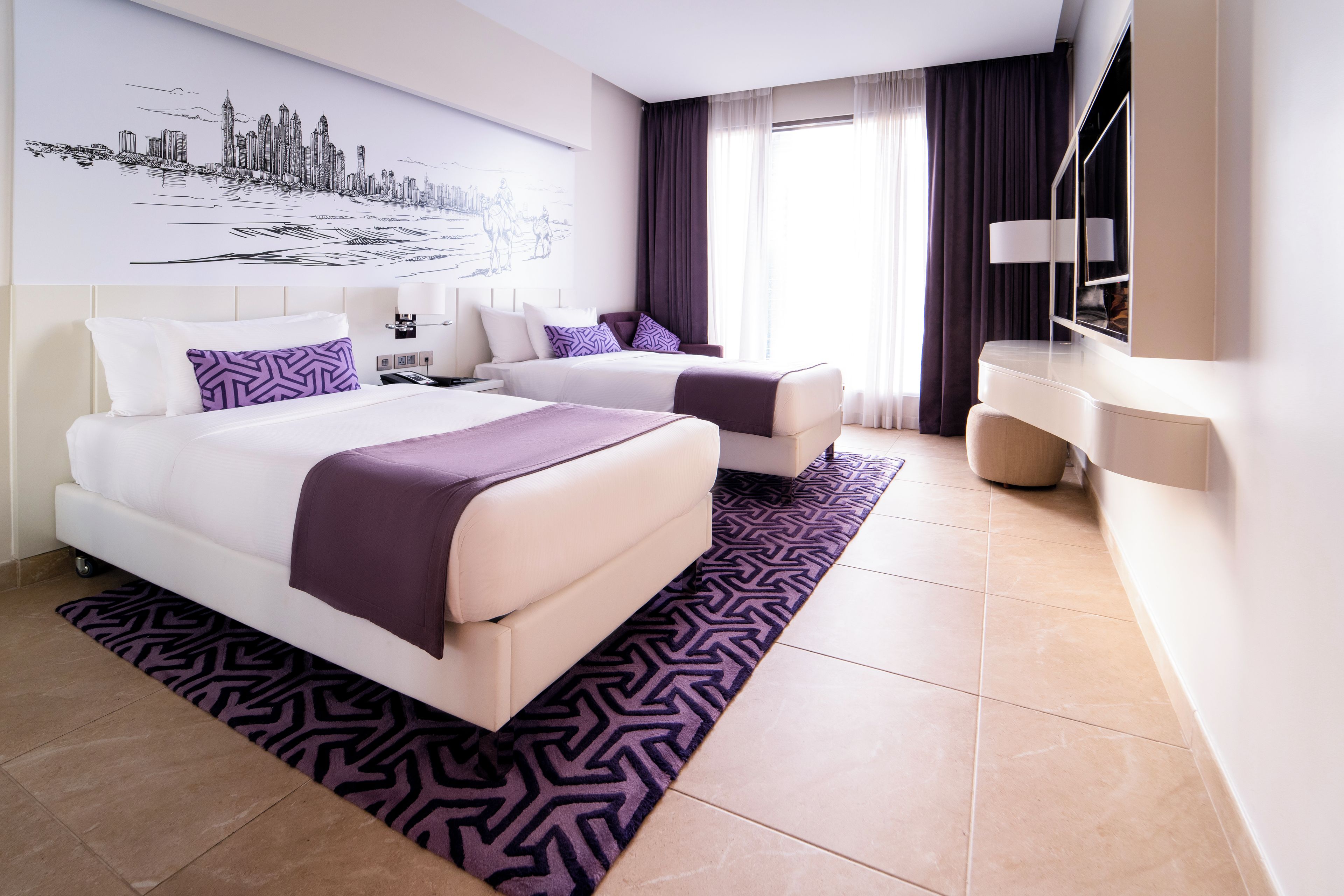 A hotel room with a large bed, a desk, and a chair. There is a window with curtains, a wall-mounted TV, and a cityscape artwork.