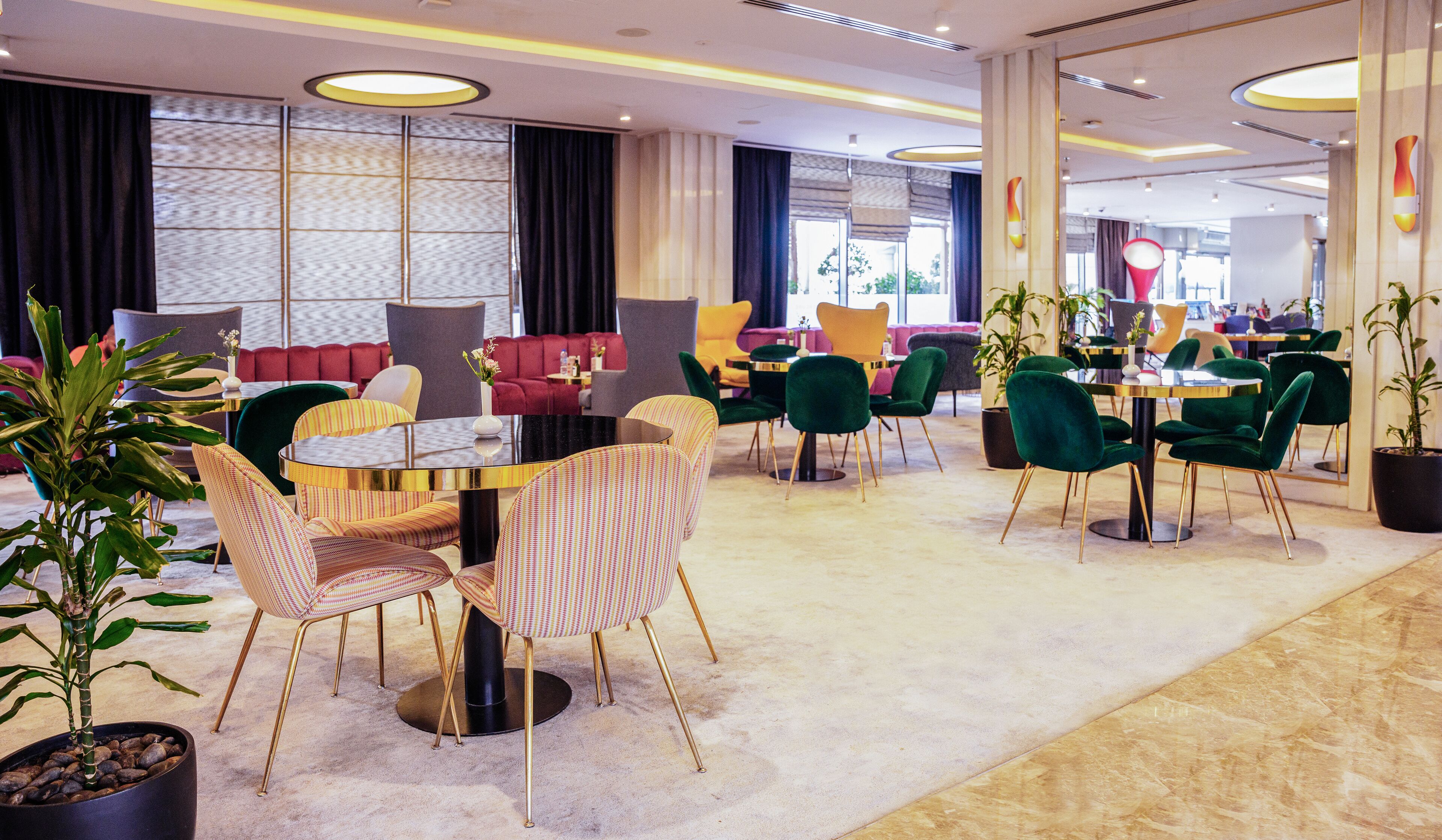 Un moderno vestíbulo de hotel con diversas zonas de estar, incluyendo una mesa redonda central con sillas, una zona de sofás y sillas individuales. Hay plantas en macetas y un gran ventanal que proporciona luz natural.